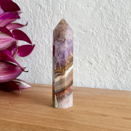 Multicolored amethyst agate crystal tower on a wooden surface with a white wall and purple flowers in the background