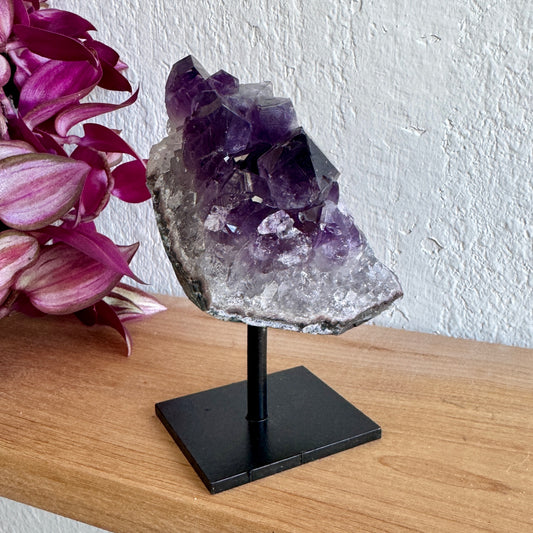 Amethyst crystal cluster on a black stand with pink flowers in the background