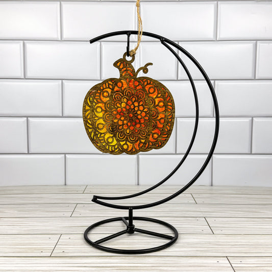 Geometric pumpkin suncatcher made from wood and acrylic hanging on a metal stand in front of a white tiled wall.