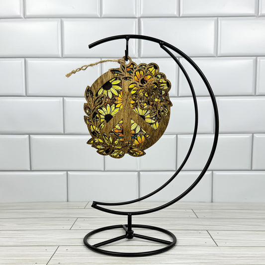 Floral peace sign suncatcher made from dark stained wood and acrylic hanging on a metal stand in front of a white tiled wall.