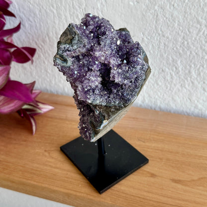 Amethyst crystal on a black stand with a white wall and pink flowers in the background