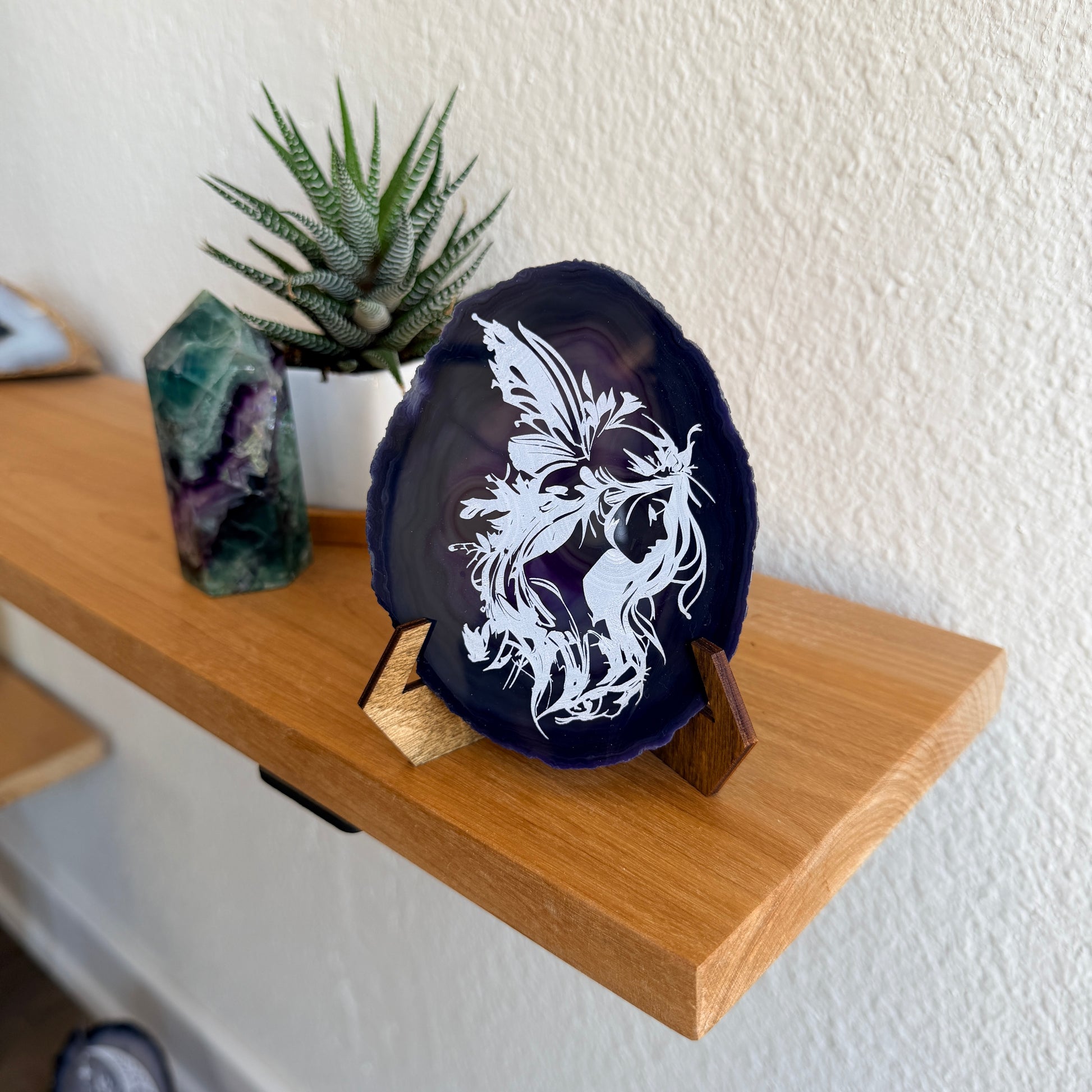 A dark purple polished agate slice with a white fairy laser engraved on it, displayed on a wooden stand on a wood shelf next to a small potted succulent and a green crystal cluster.