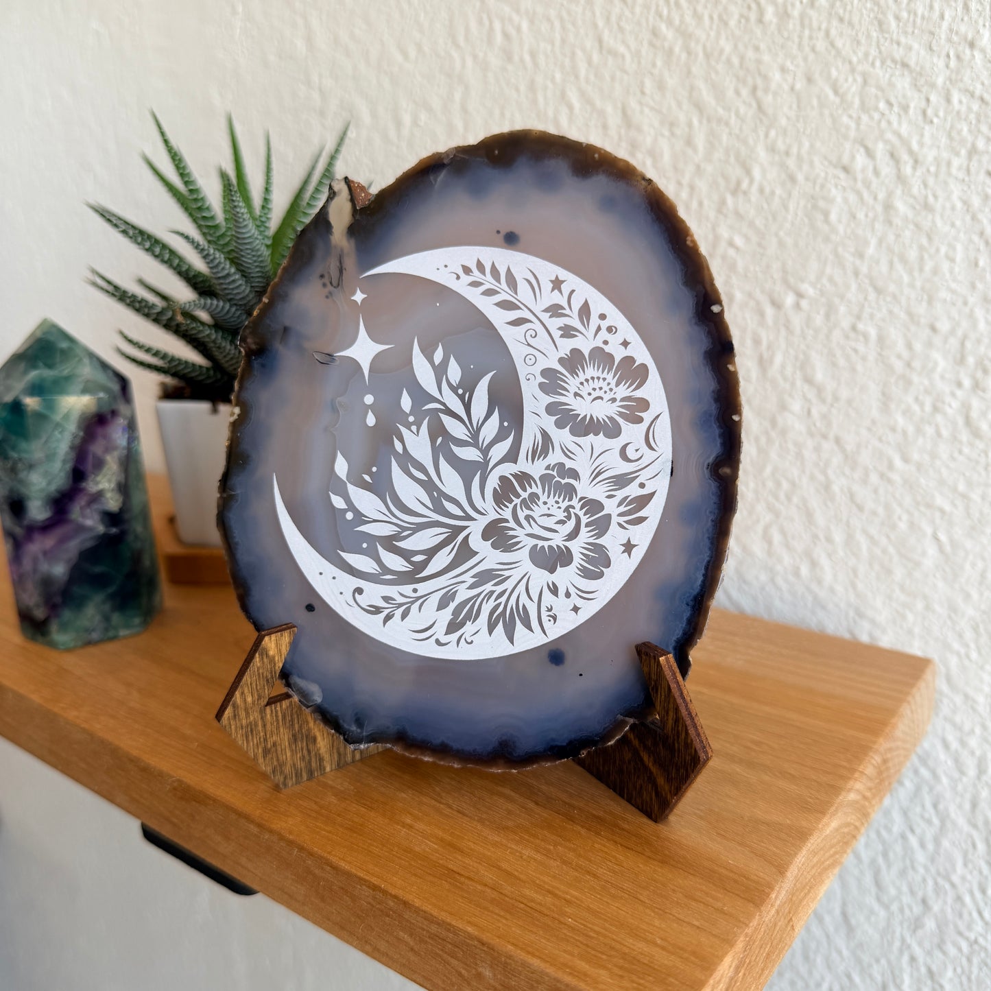 A decorative agate slice with a white laser engraved crescent moon and floral design, displayed on a small wooden stand on a wooden shelf.