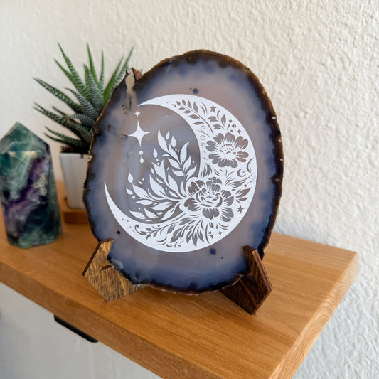 A decorative agate slice with a white laser engraved crescent moon and floral design, displayed on a small wooden stand on a wooden shelf.