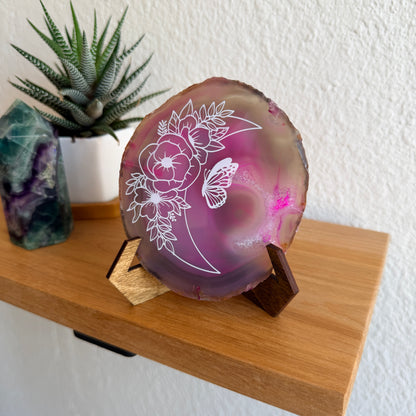 A polished agate crystal slice with white floral and moon engravings, displayed on a small wooden stand on a wooden shelf next to a potted succulent.