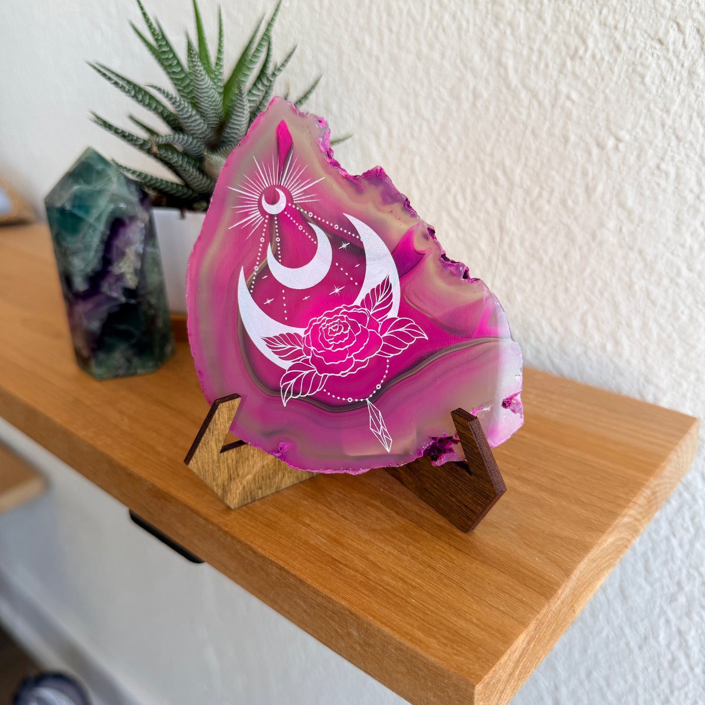 A pink agate crystal slice with white crescent moon and rose design laser engraved on it, displayed on a wooden stand on a wooden shelf next to a crystal cluster and a small plant.