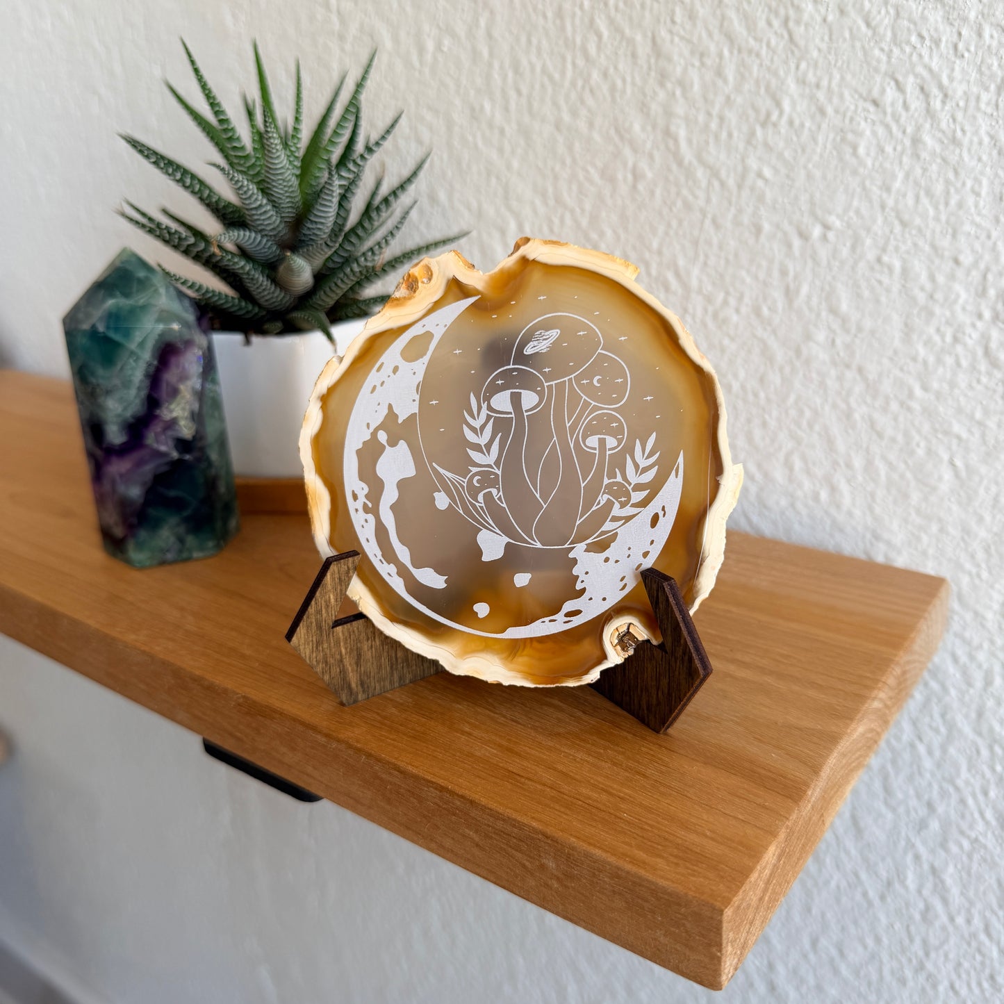 A decorative mushroom and moon engraved natural agate slice, displayed on a small wooden stand on a wooden shelf next to a potted succulent and a purple crystal.