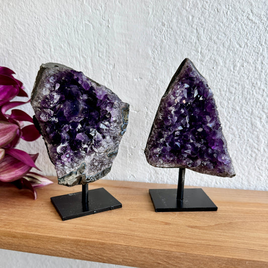 Two amethyst crystal specimens on stands with a white wall background