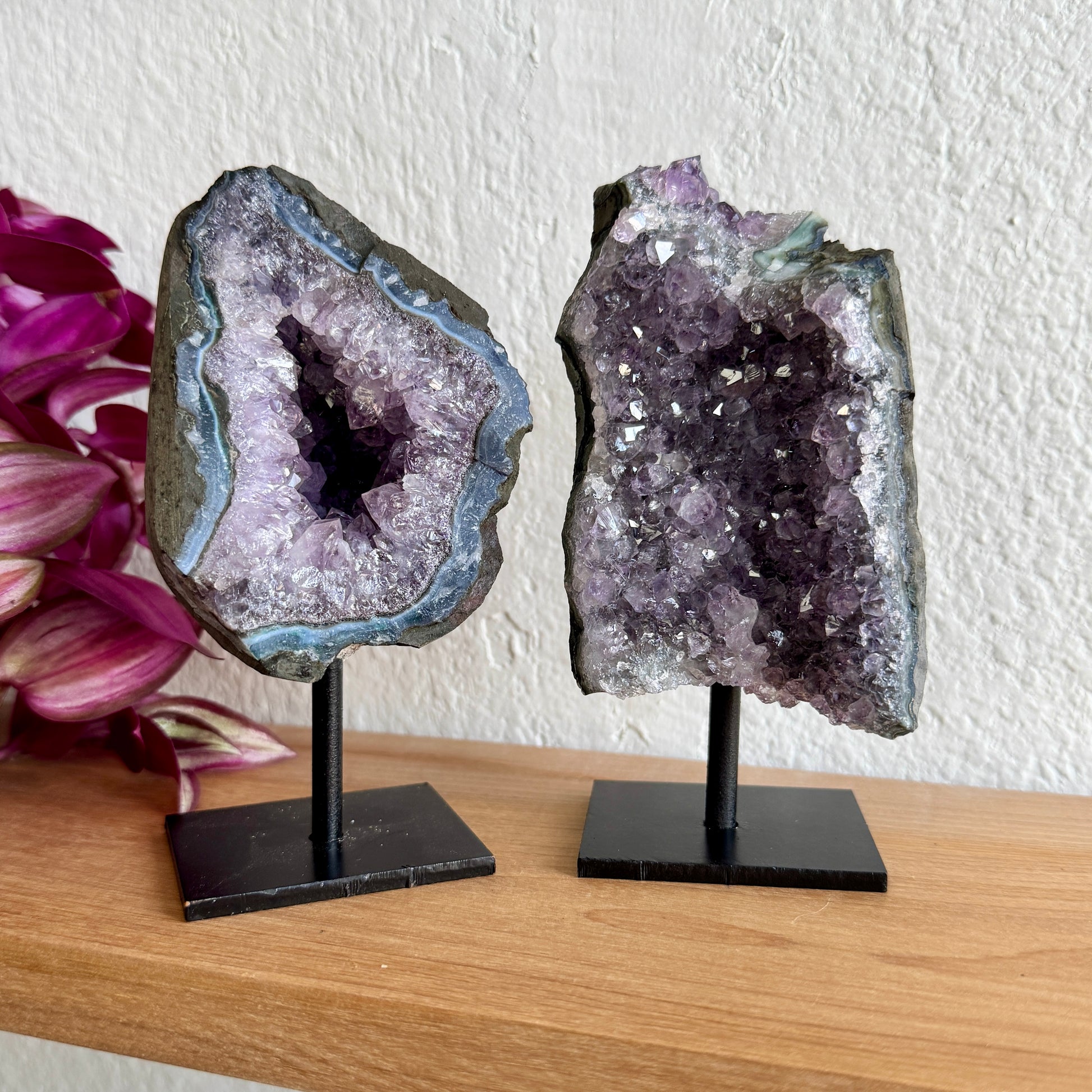 Two amethyst geode crystals on stands with a white wall and pink flowers in the background.