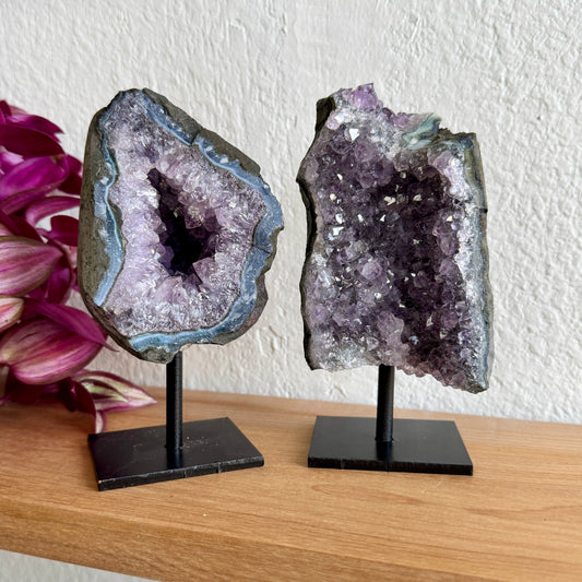 Two amethyst geode crystals on stands with a white wall and pink flowers in the background.