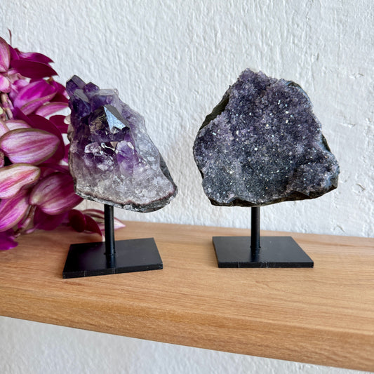 Two amethyst crystal specimens on stands with a white wall and pink flowers in the background