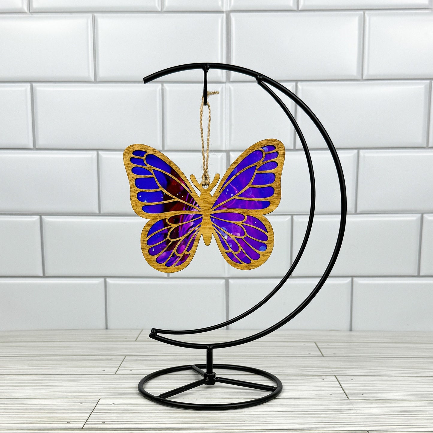 Butterfly suncatcher made from wood and acrylic shown in light stain option hanging on a metal stand in front of a white tiled wall.