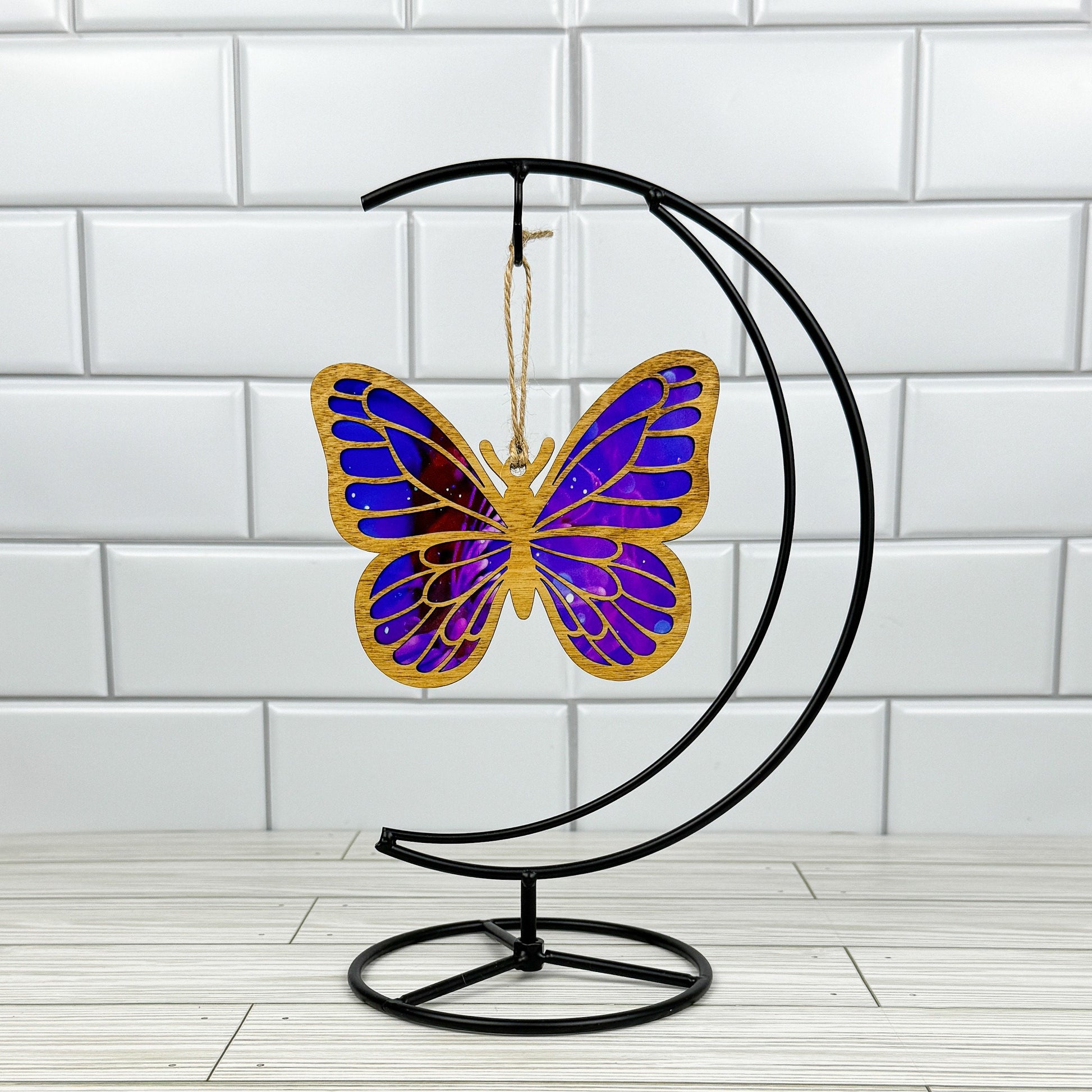 Butterfly suncatcher made from wood and acrylic shown in light stain option hanging on a metal stand in front of a white tiled wall.