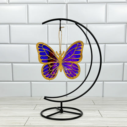 Butterfly suncatcher made from wood and acrylic shown in light stain option hanging on a metal stand in front of a white tiled wall.