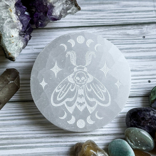 Selenite stone with engraved death head moth design on a wooden surface with crystals around