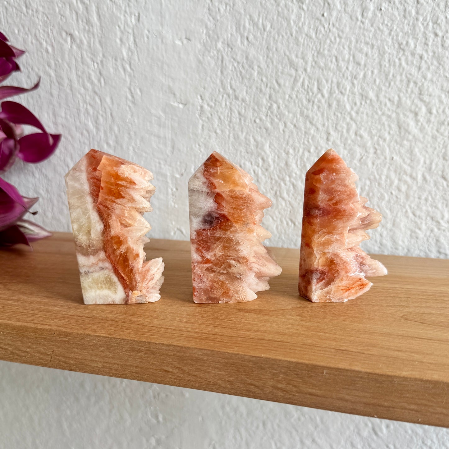 Three orange dogtooth calcite crystal towers on a wooden shelf with a white wall background