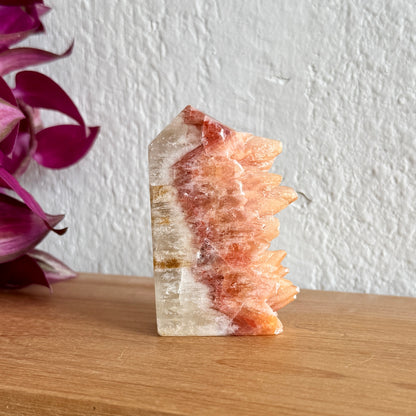 Dogtooth calcite crystal tower on a wooden surface with a white wall and purple plant in the background