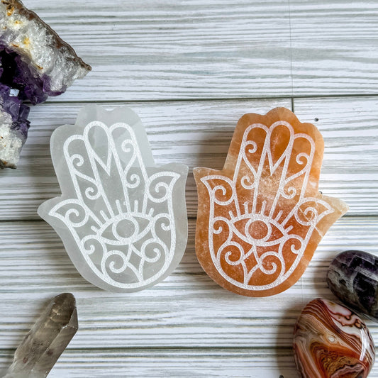Two engraved selenite Hamsa hand-shaped stones on a wooden surface with crystals around.