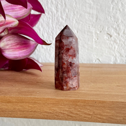 Hematoid quartz crystal tower on a wooden surface with pink flowers in the background
