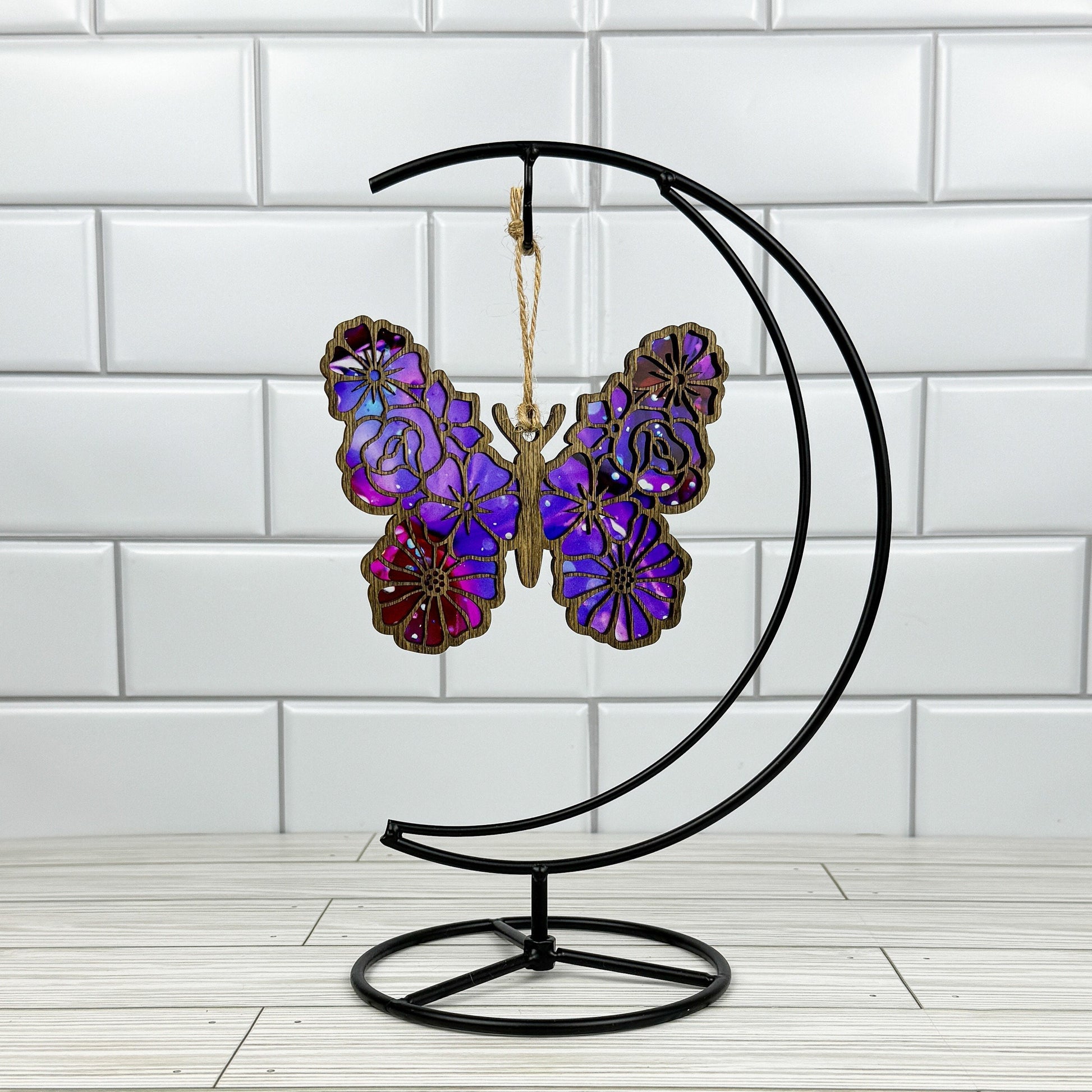 Floral butterfly suncatcher made from wood and acrylic shown in dark stain option hanging on a metal stand in front of a white tiled wall.