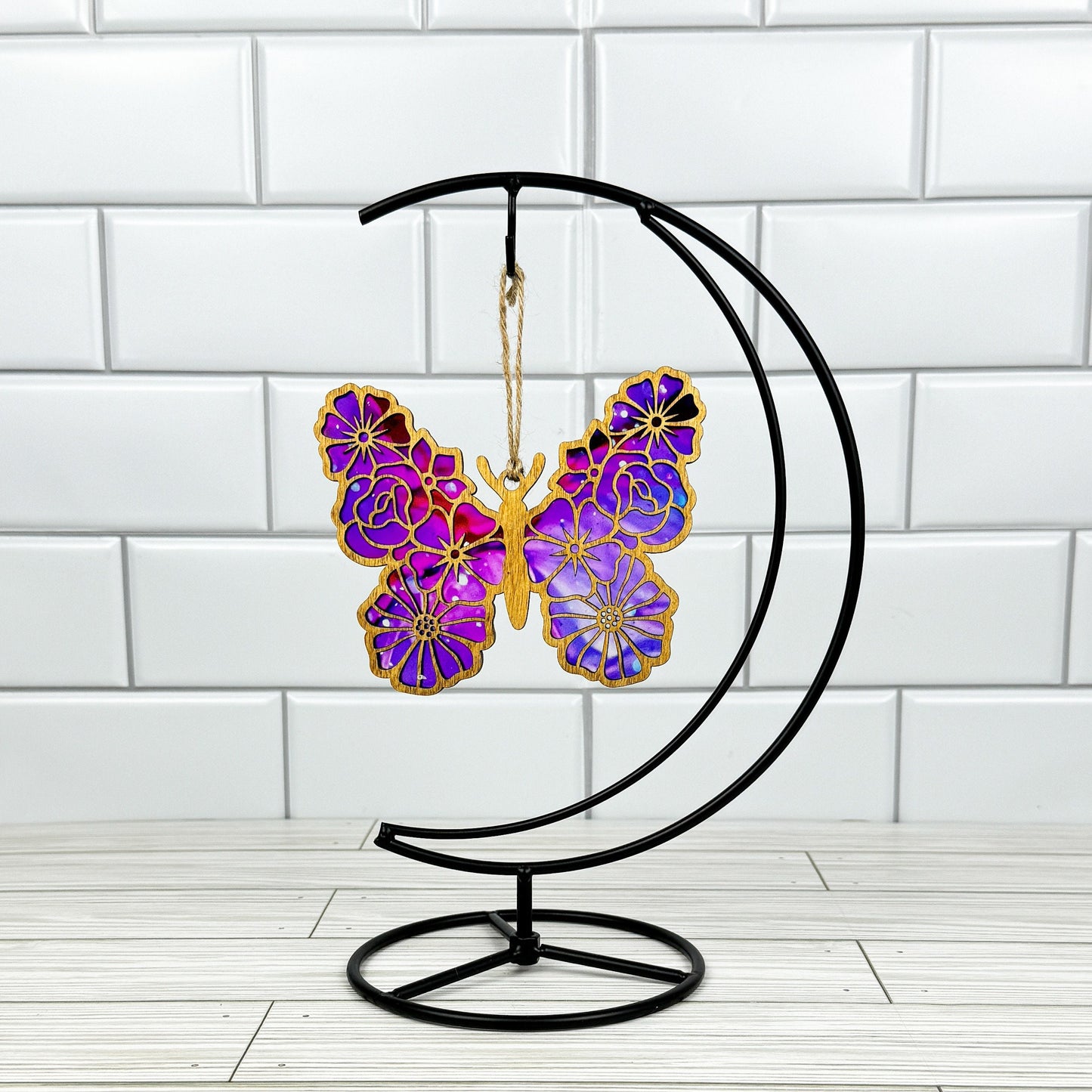 Floral butterfly suncatcher made from wood and acrylic shown in light stain option hanging on a metal stand in front of a white tiled wall.