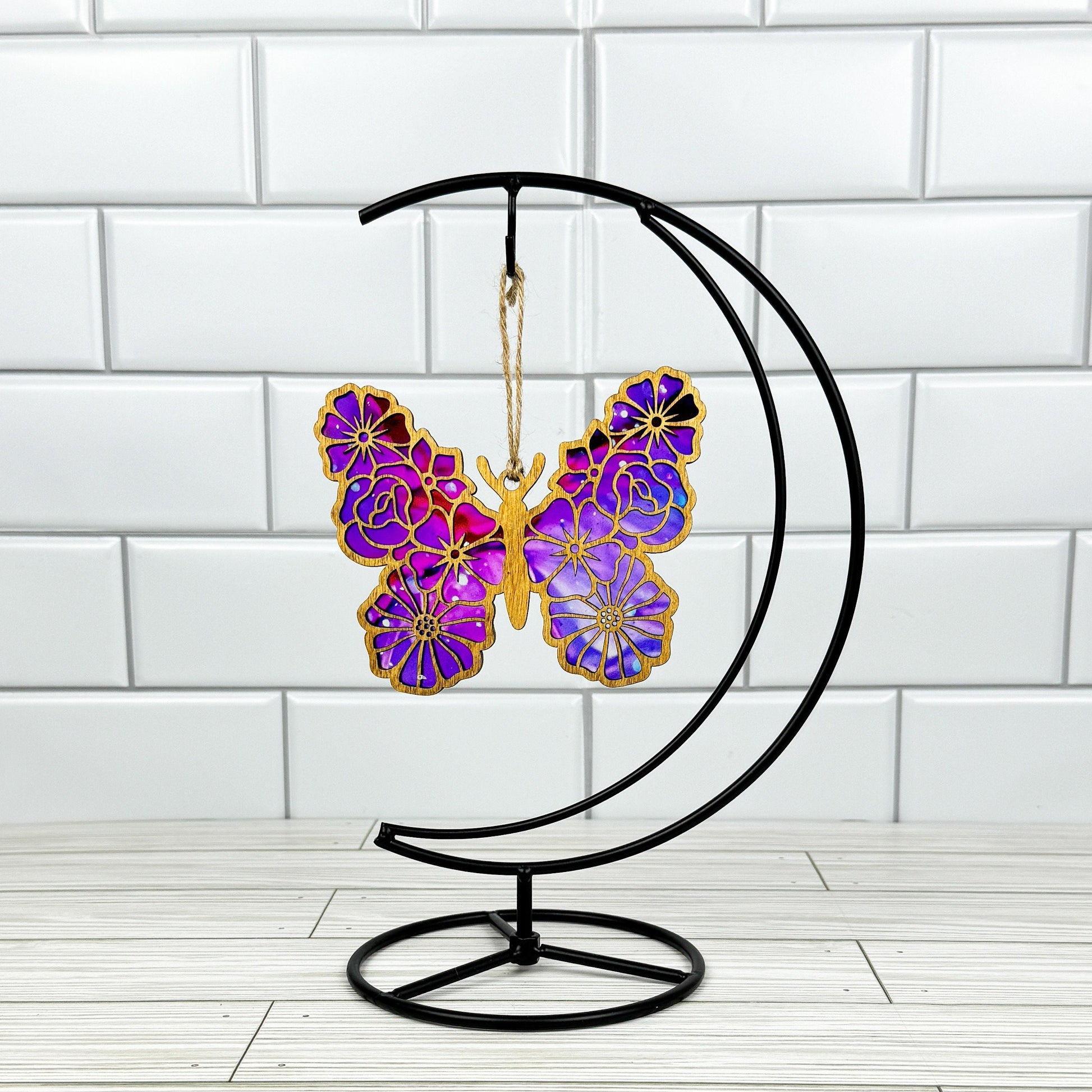 Floral butterfly suncatcher made from wood and acrylic shown in light stain option hanging on a metal stand in front of a white tiled wall.