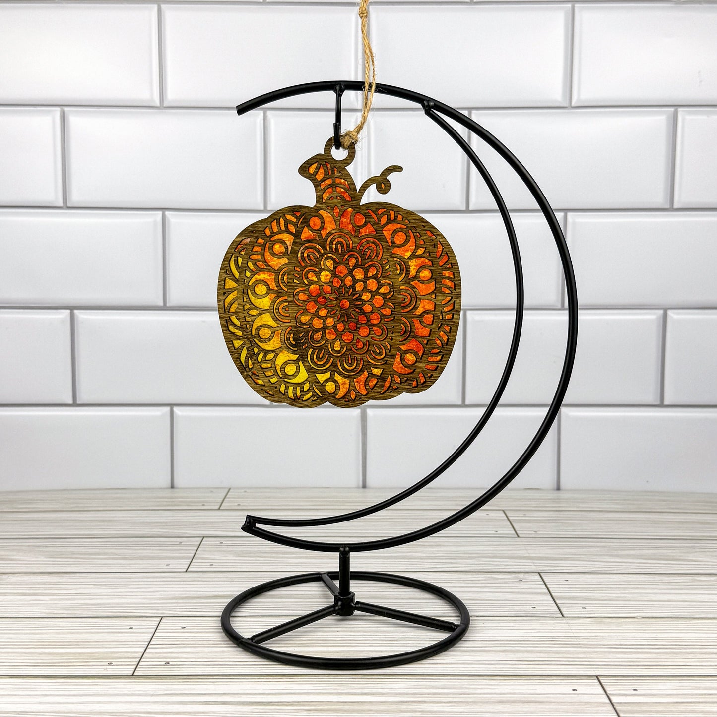 Geometric pumpkin suncatcher made from wood and acrylic hanging on a metal stand in front of a white tiled wall.