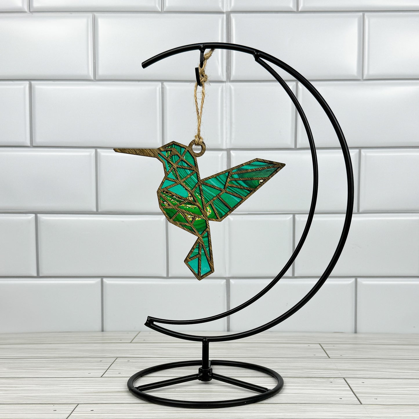 Hummingbird suncatcher made from wood and acrylic hanging on a metal stand in front of a white tiled wall.
