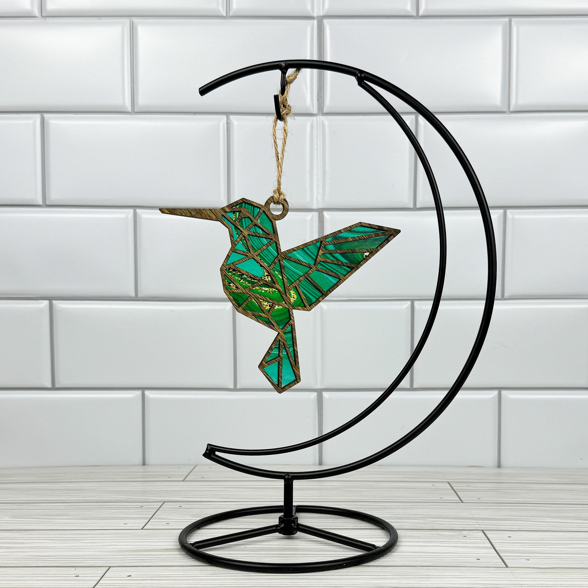 Hummingbird suncatcher made from wood and acrylic hanging on a metal stand in front of a white tiled wall.