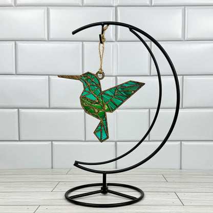 Hummingbird suncatcher made from wood and acrylic hanging on a metal stand in front of a white tiled wall.