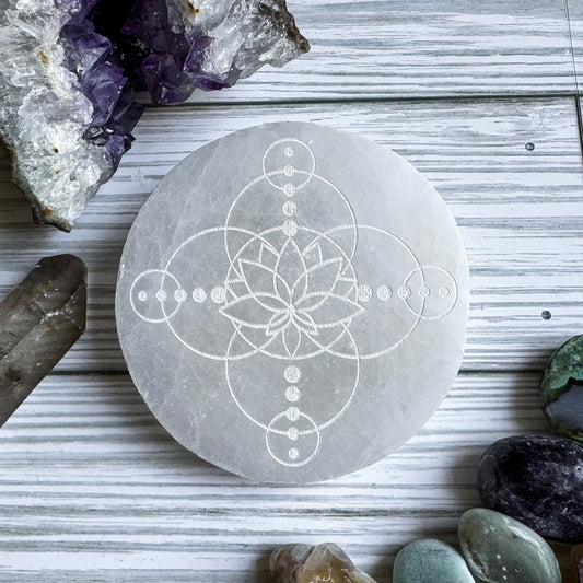 Selenite with a geometric lotus flower design on a wooden surface surrounded by crystals and stones.