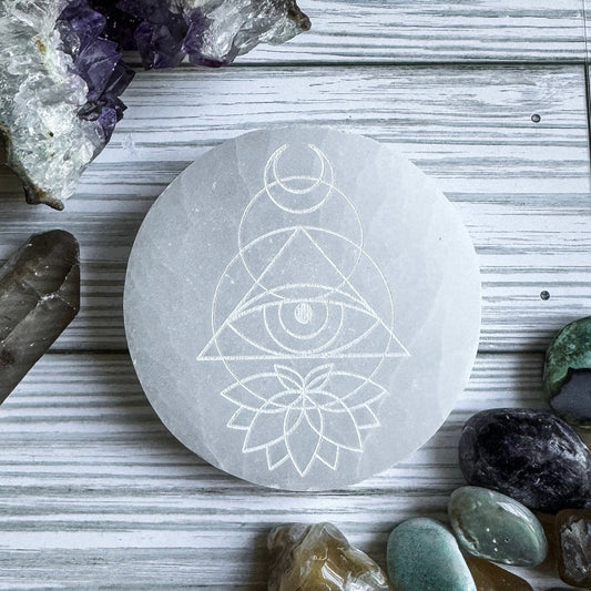 Selenite stone with engraved lotus and eye geometric design surrounded by crystals on a wooden surface