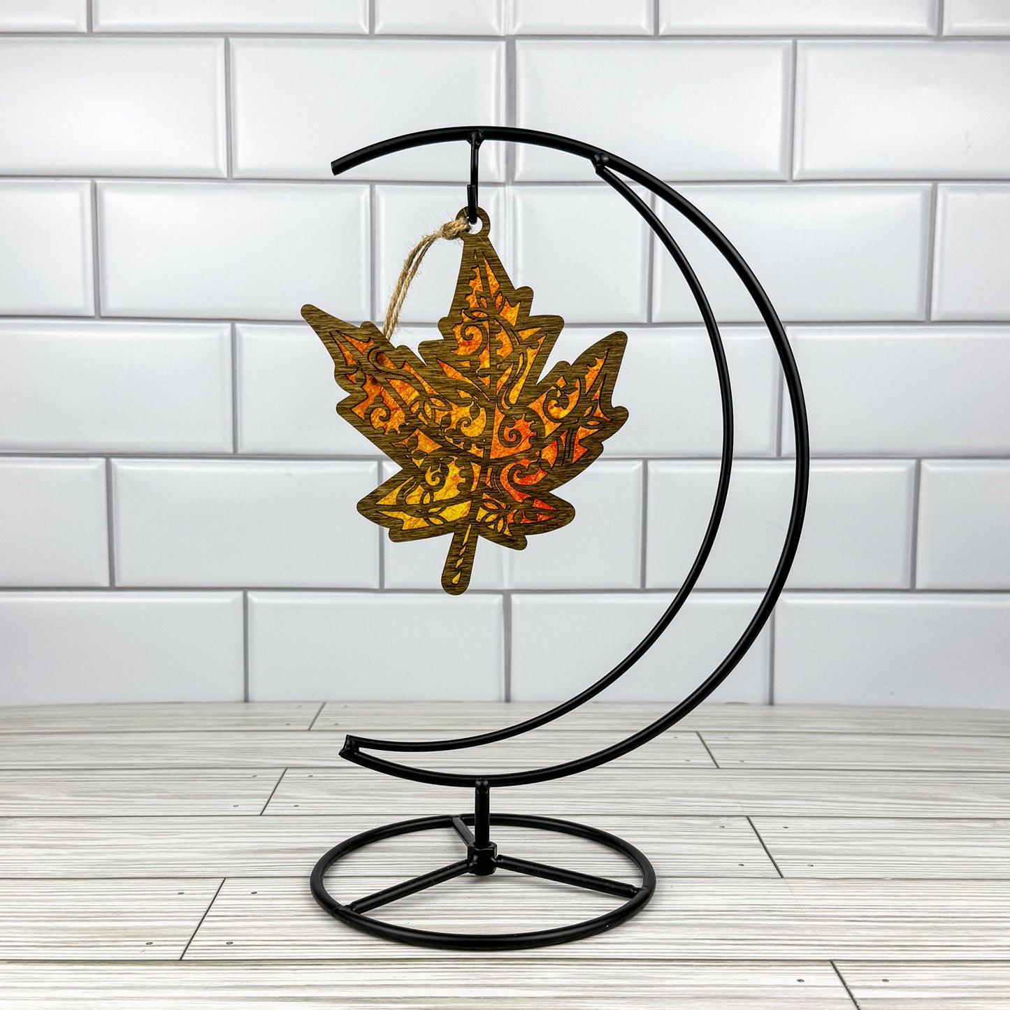 Maple leaf suncatcher made from wood and acrylic hanging on a metal stand in front of a white tiled wall.