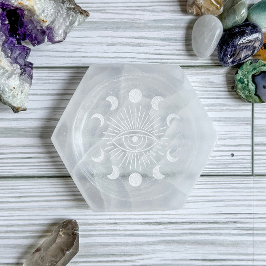 Selenite hexagonal crystal dish with engraved moon and eye design on a wooden surface with crystals around