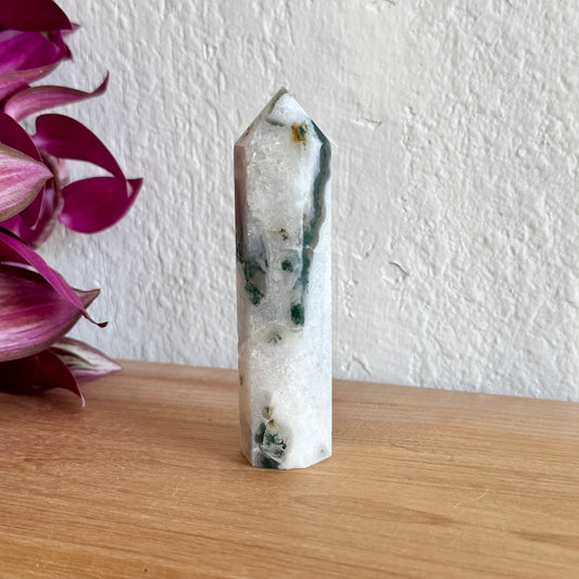 Moss agate crystal tower on a wooden surface with a white wall and purple flowers in the background