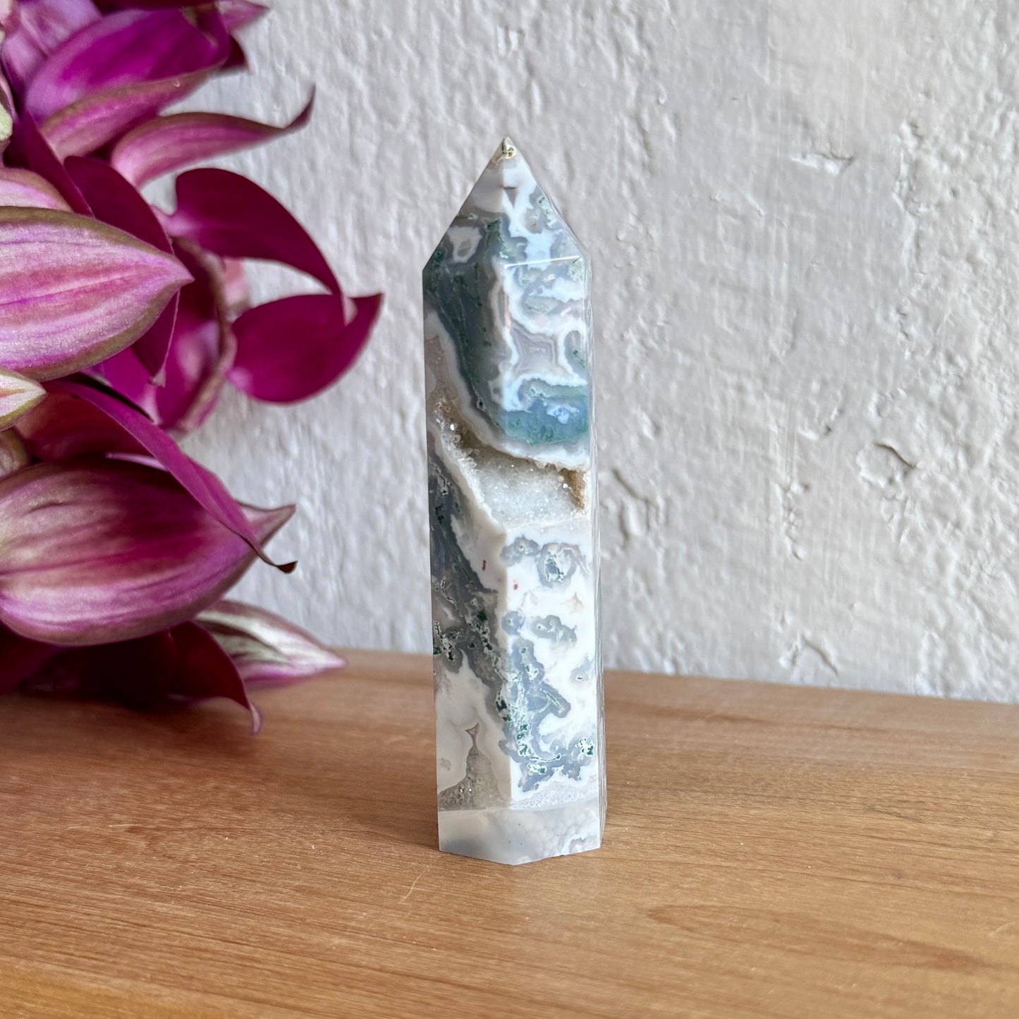Moss agate crystal tower on a wooden surface with pink flowers in the background