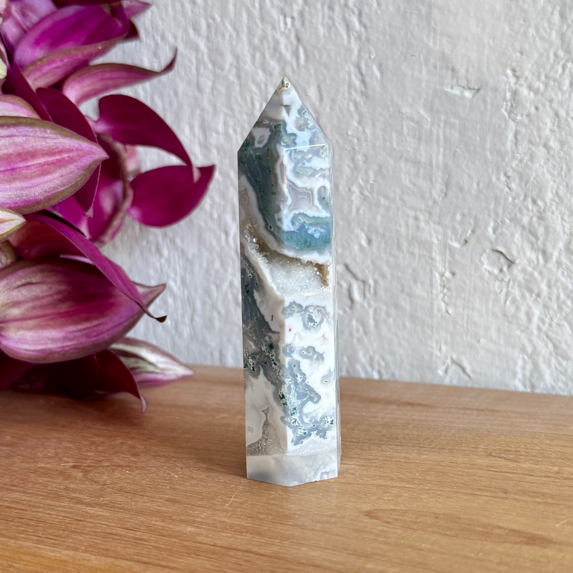 Moss agate crystal tower on a wooden surface with pink flowers in the background