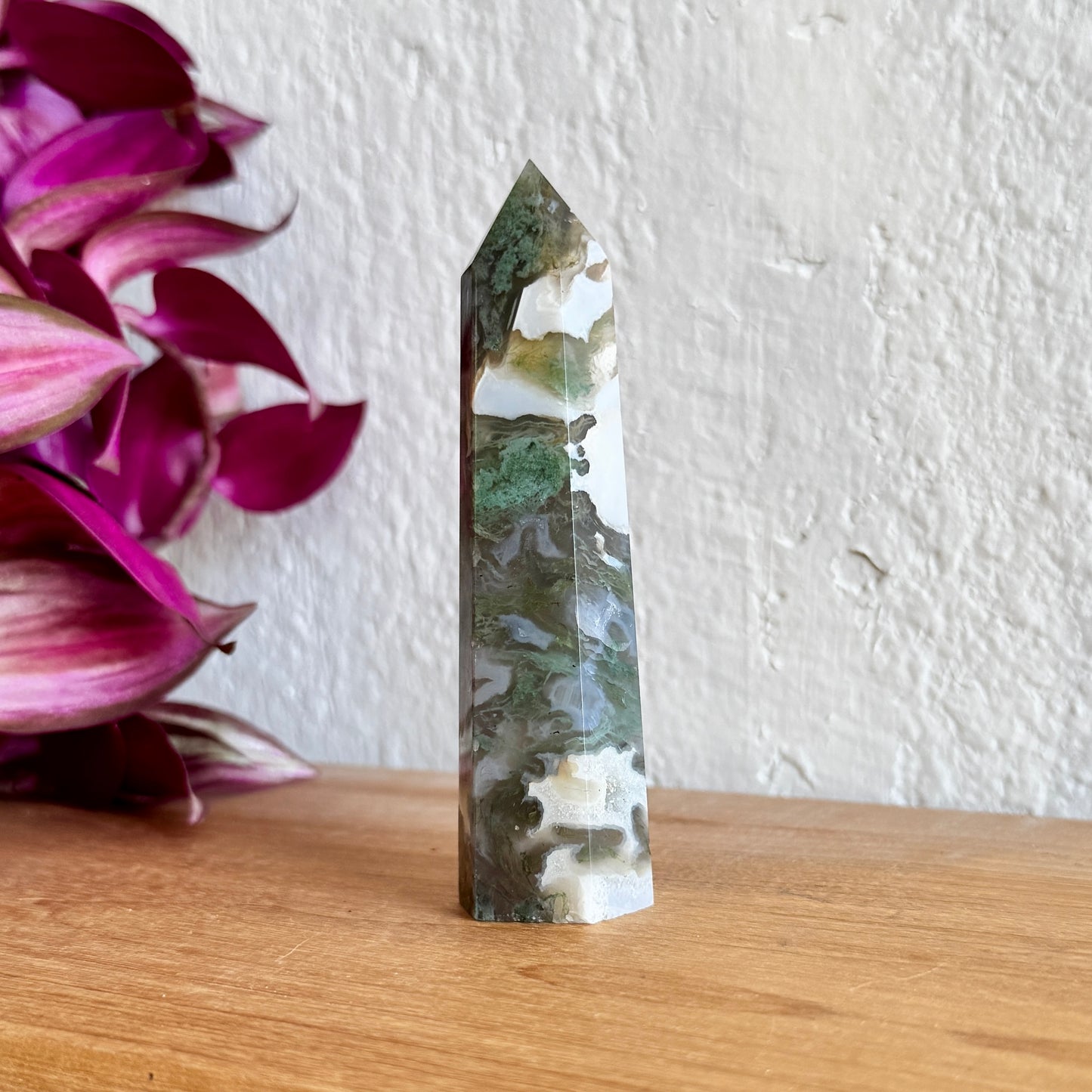 One moss agate crystal tower on a wooden surface with a white wall background
