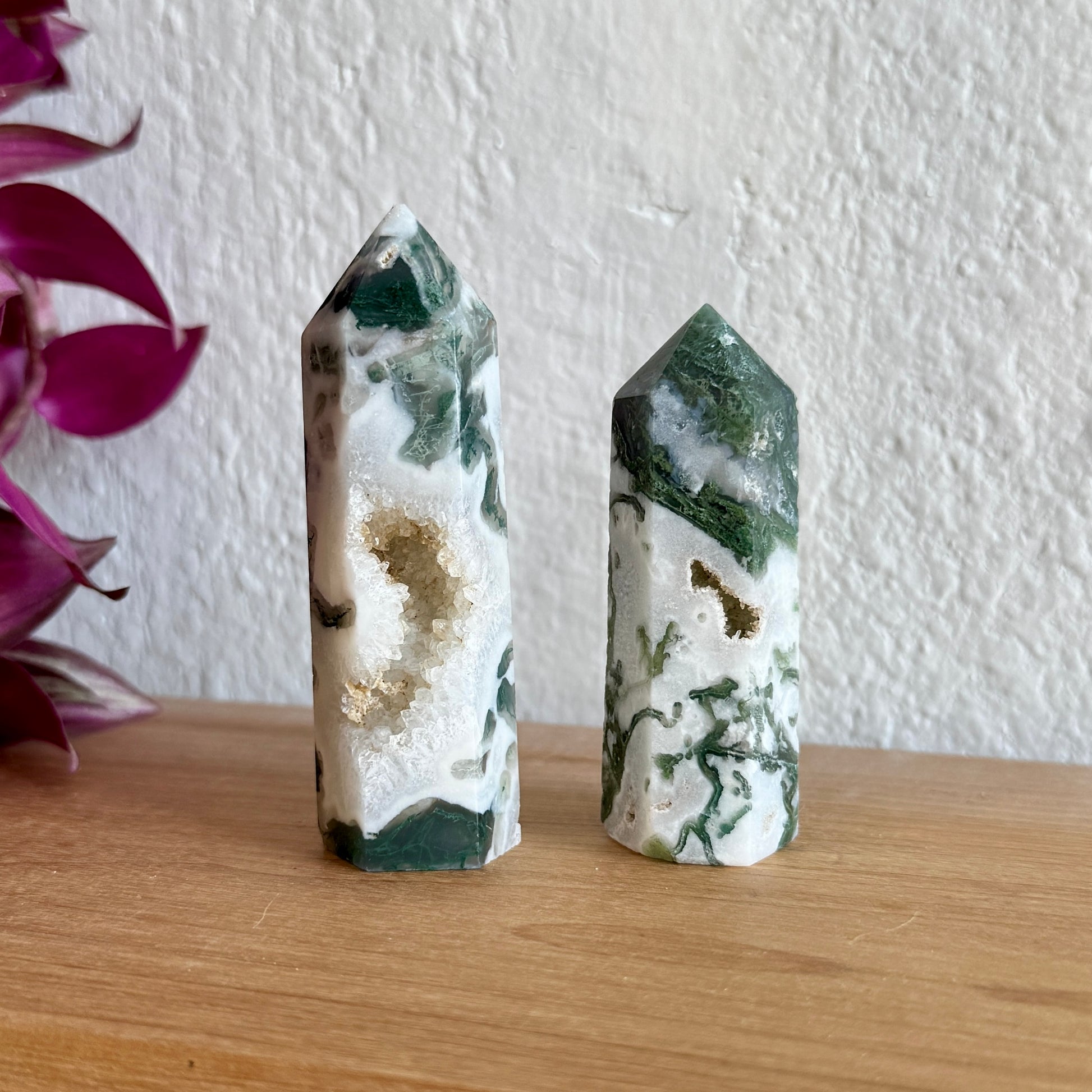 Two moss agate crystal towers on a wooden surface with a white wall background