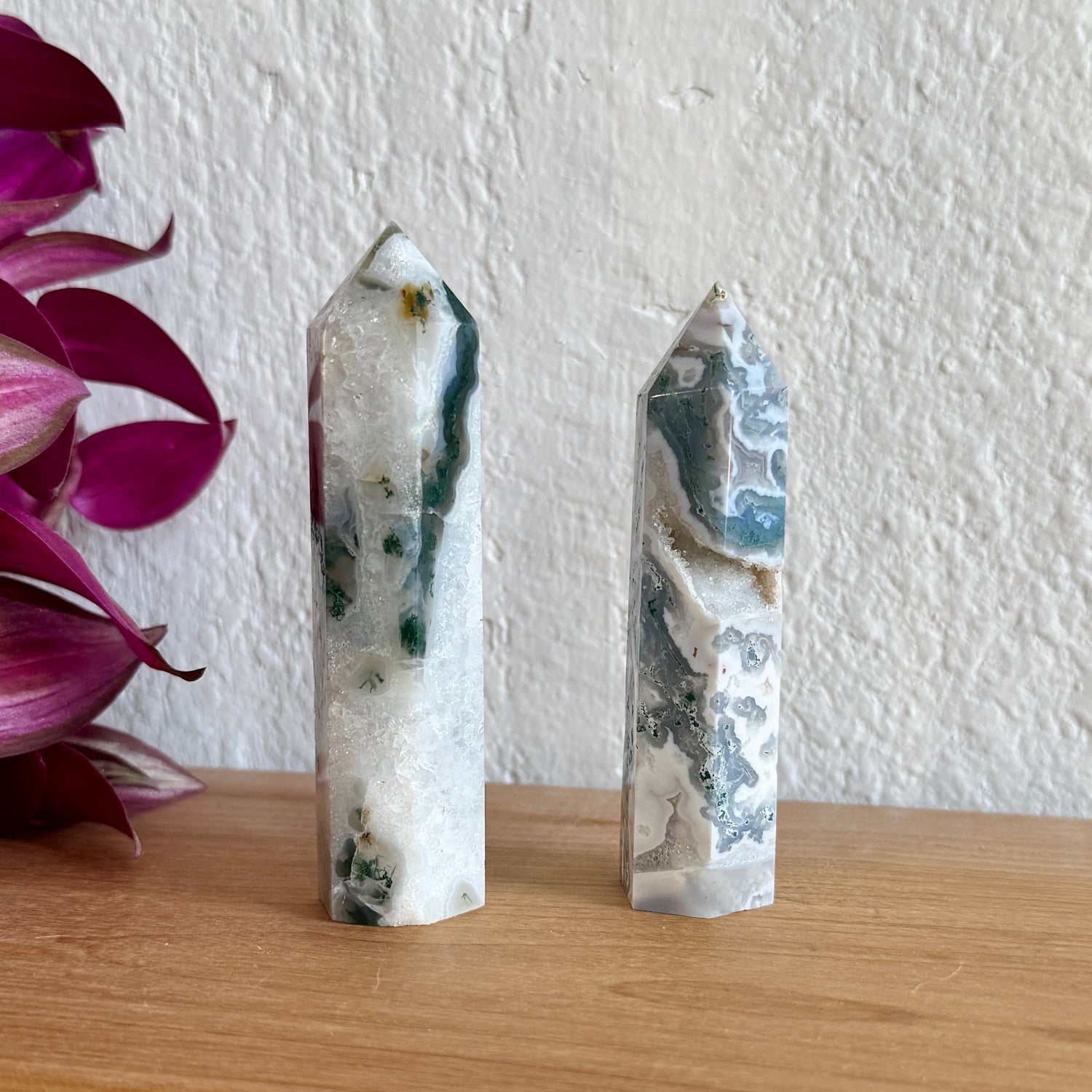 Two moss agate crystal towers on a wooden surface with a white wall and purple flowers in the background.