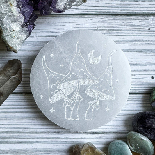 Selenite stone with engraved design of mushrooms and moon on a wooden surface with crystals.