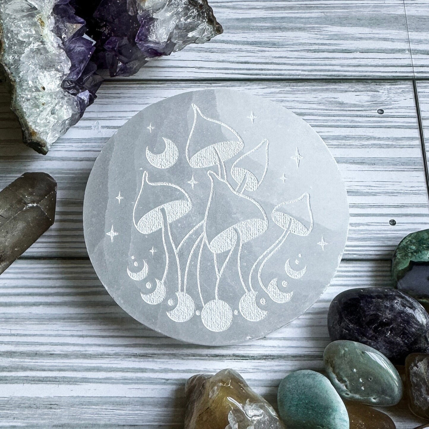 Selenite stone with engraved mushroom and moon design on a wooden surface with crystals around