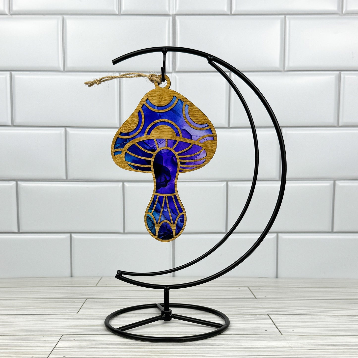 Mushroom suncatcher made from wood and acrylic shown in the blue and purple option hanging on a metal stand in front of a white tiled wall.
