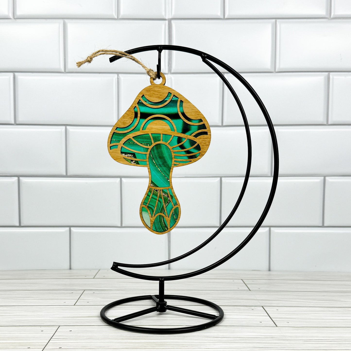 Mushroom suncatcher made from wood and acrylic shown in the green option hanging on a metal stand in front of a white tiled wall.