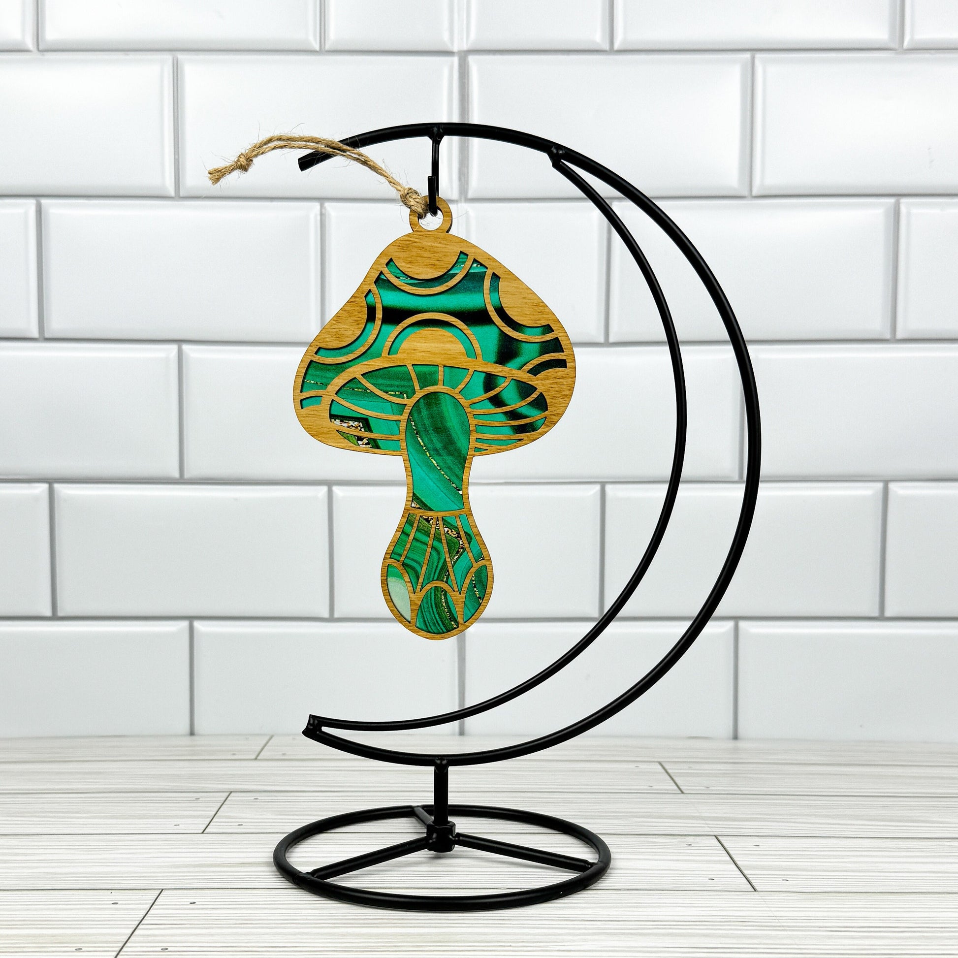 Mushroom suncatcher made from wood and acrylic shown in the green option hanging on a metal stand in front of a white tiled wall.