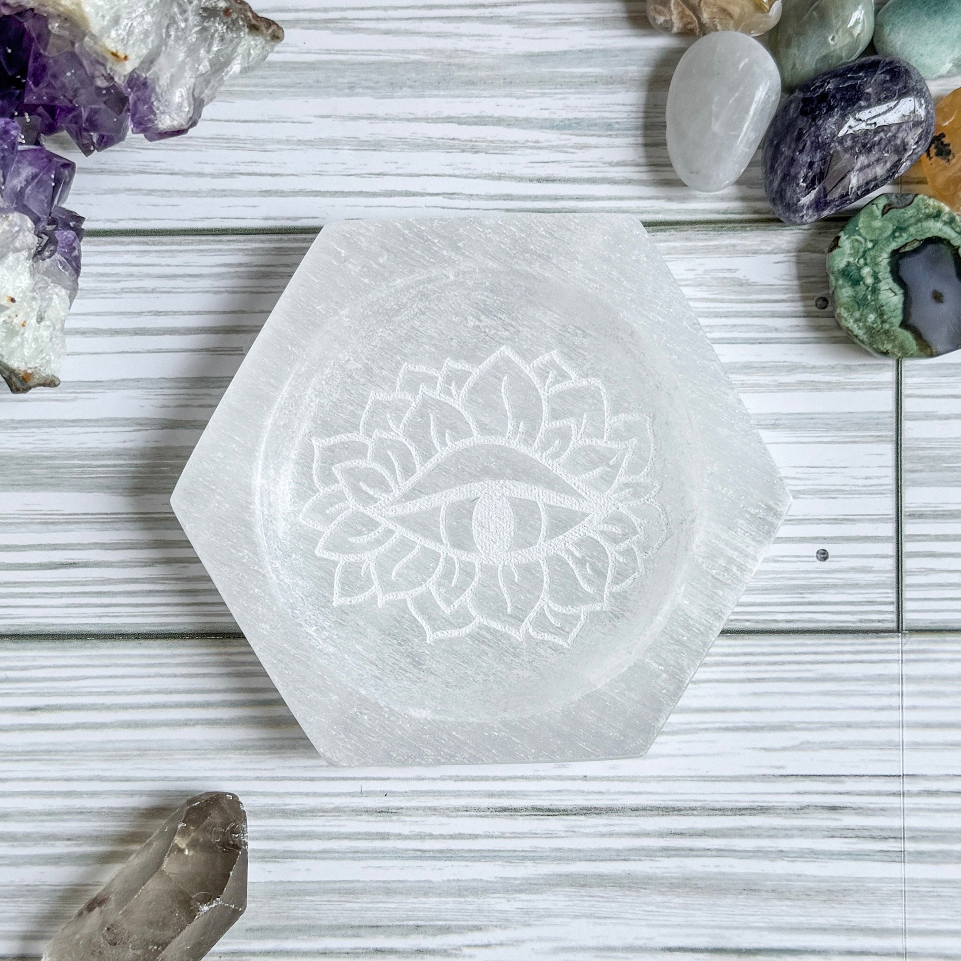 Hexagonal selenite crystal dish with mystical eye engraving on a wooden surface with various crystals around