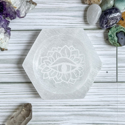 Hexagonal selenite crystal dish with mystical eye engraving on a wooden surface with various crystals around