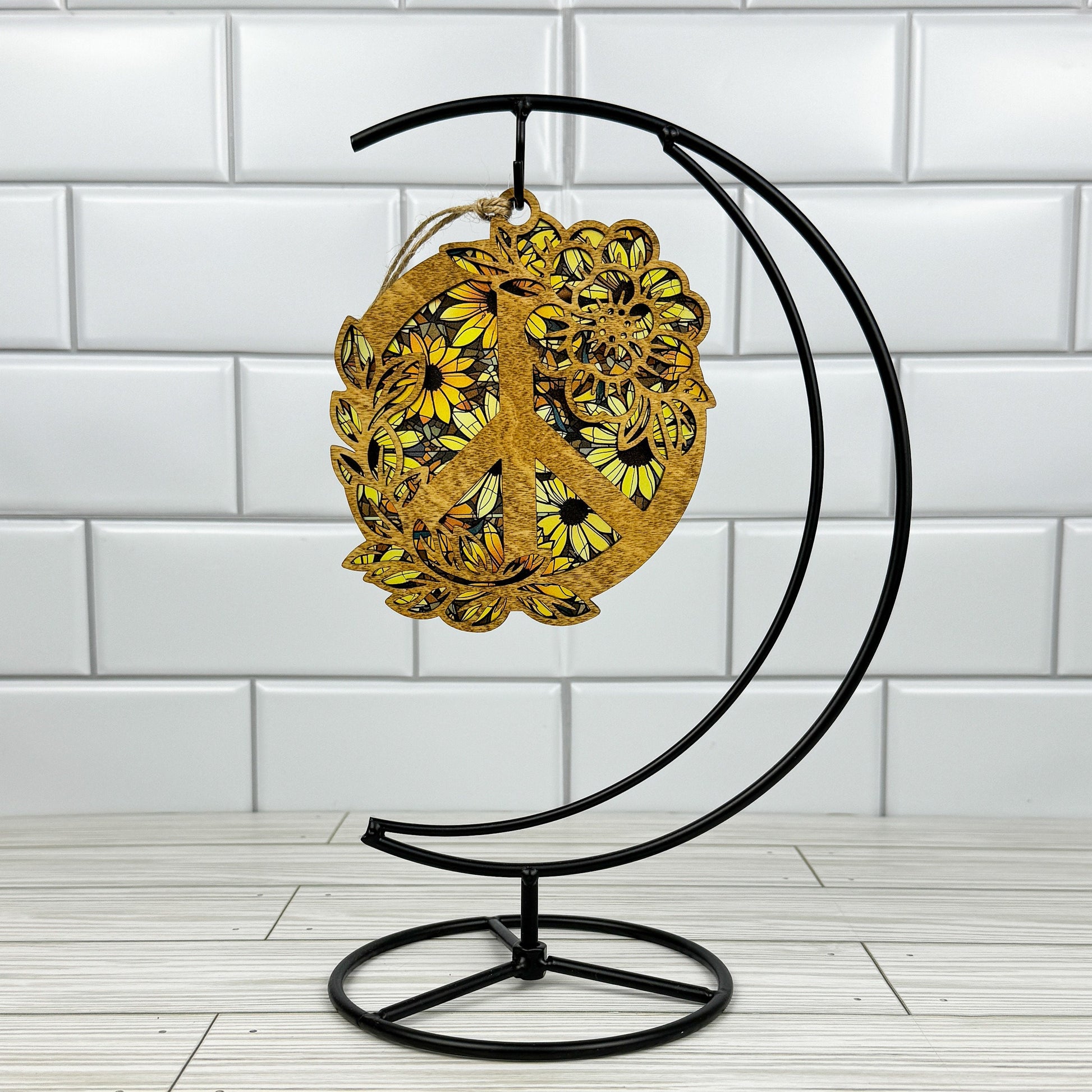 Floral peace sign suncatcher made from light stained wood and acrylic hanging on a metal stand in front of a white tiled wall.