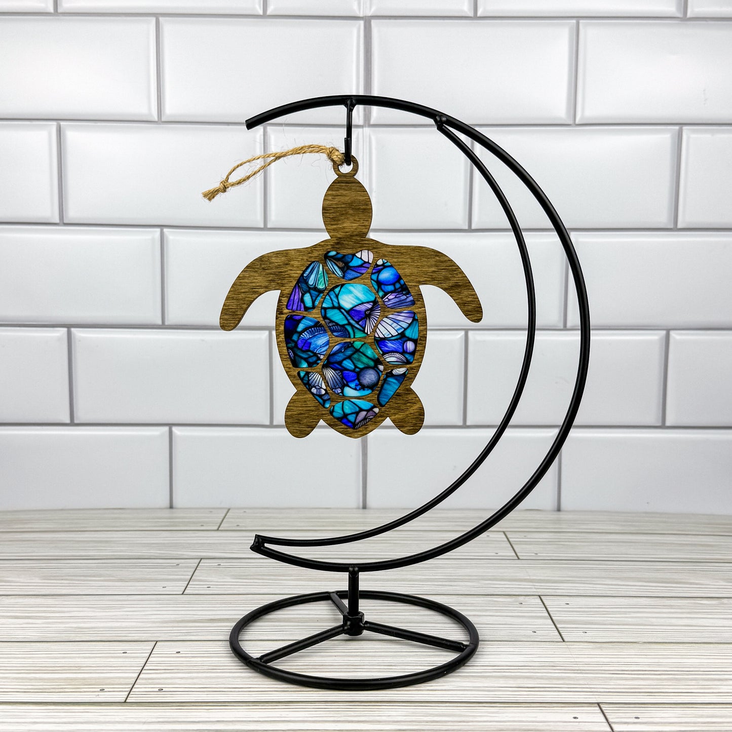 Sea turtle suncatcher made from wood and acrylic hanging on a metal stand in front of a white tiled wall.