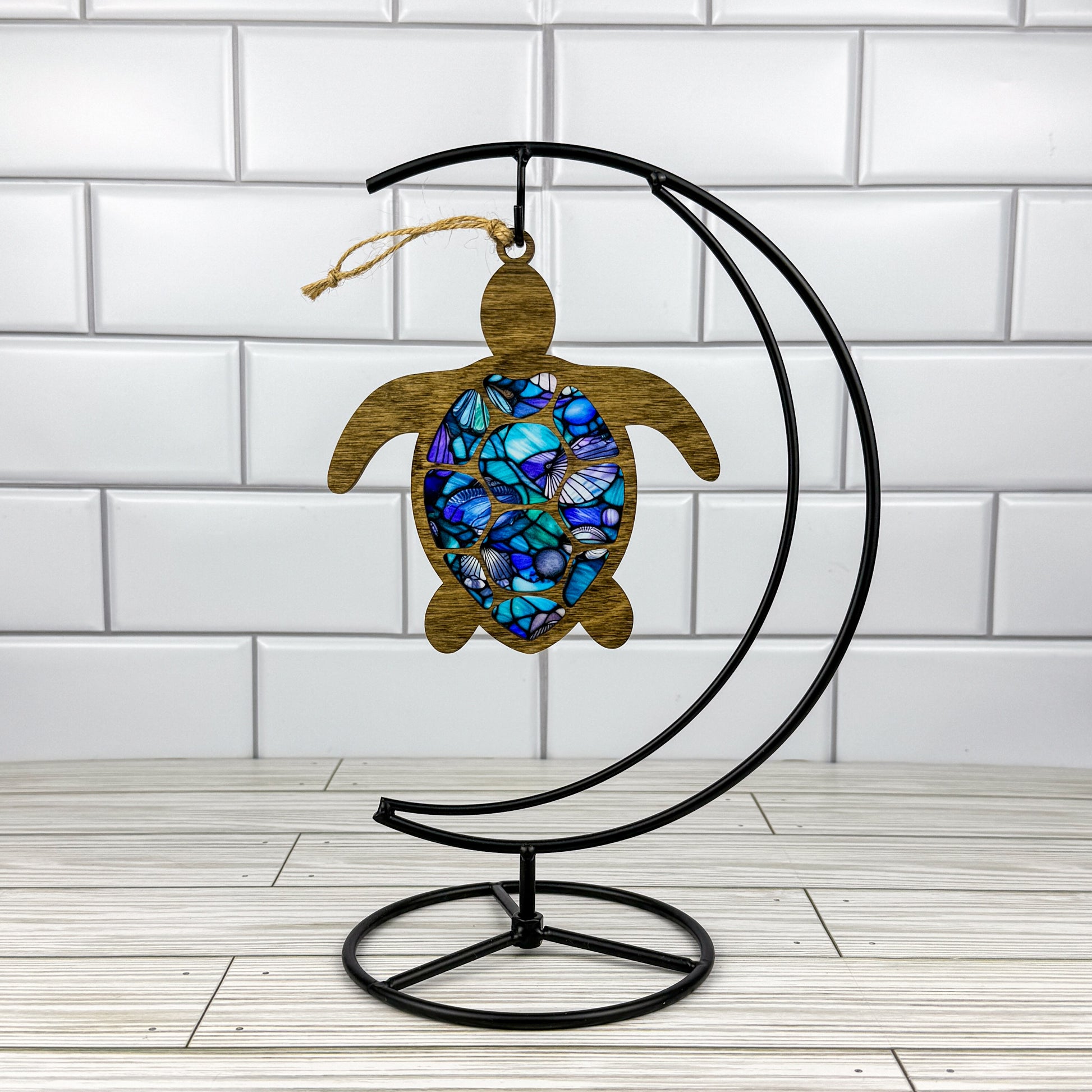 Sea turtle suncatcher made from wood and acrylic hanging on a metal stand in front of a white tiled wall.